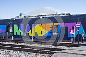 A colorful wall mural with the word âMariettaâ along the railroad tracks with people standing on the corner in Marietta